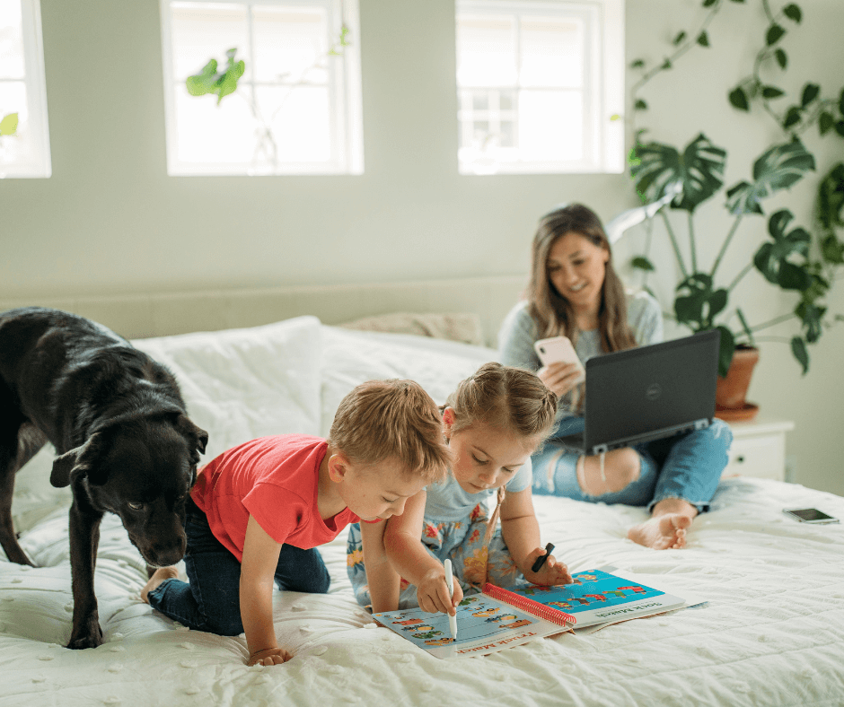 Two children reading a book kneeling on a bed with a black Labrador and female adult on a laptop.